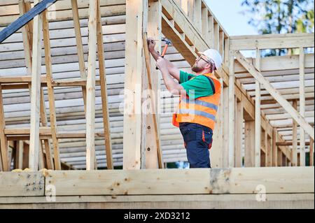 Charpentier construisant une maison à ossature en bois de deux étages près de la forêt. Homme barbu dans des lunettes martelant des clous dans la structure tout en portant casque de protection et gilet de construction. Banque D'Images