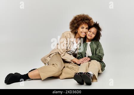 Mère afro-américaine bouclée et fille dans une tenue élégante, assis sur le sol, souriant chaleureusement l'un à l'autre. Banque D'Images