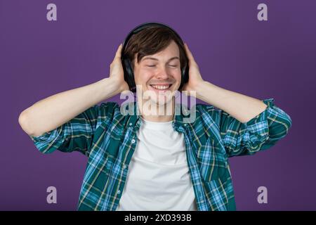 Jeune homme dans une chemise à carreaux vert souriant et appréciant la musique avec des écouteurs sur un fond violet. Guy écoute ses morceaux préférés, en appuyant les écouteurs sur ses oreilles avec ses mains. Banque D'Images