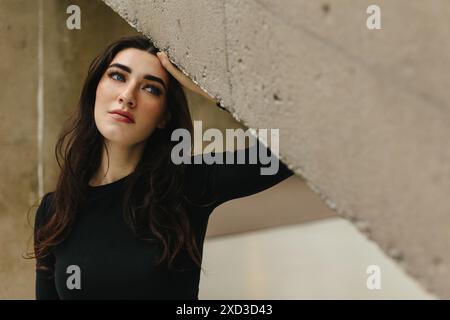 Portrait d'une jeune femme avec une expression contemplative penchée sous un élément architectural concret, mettant en valeur ses cheveux foncés et son makeu vibrant Banque D'Images