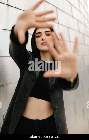 Une jeune femme pose expressivement dans un environnement urbain, en mettant l'accent sur ses mains encadrant son visage contre un mur gris pâle Banque D'Images