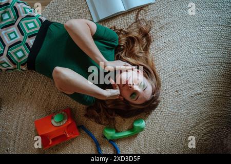 Vue de dessus d'une jeune femme avec les yeux fermés dans une robe rétro verte se trouve sur un tapis tissé, tenant ludique un téléphone, évoquant la scène de femme au foyer des années 1950. Banque D'Images