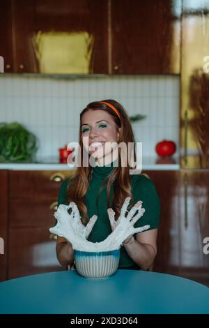 Une femme caucasienne rousse heureuse vêtue d'une tenue des années 50, s'engageant ludique avec de la pâte dans une cuisine de style rétro, regarde directement la caméra avec joie Banque D'Images