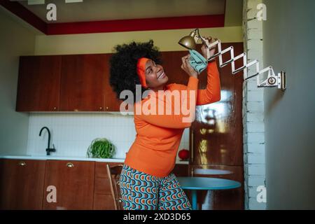 Femme noire afro-américaine ludique dans une tenue vibrante des années 50 équilibrant une lampe de table, dans un cadre de cuisine rétro, regardant la lampe avec une joie Banque D'Images