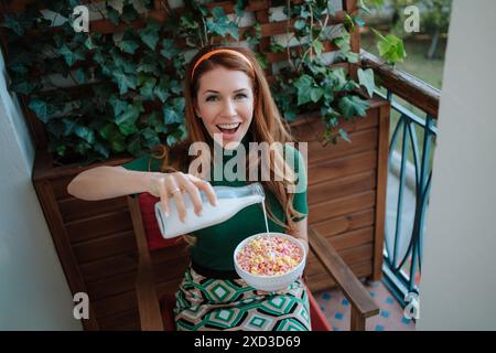 Une joyeuse femme rousse, vêtue d'une tenue vibrante de style années 1950, verse du lait dans un bol de céréales colorées tout en étant assise sur un balcon confortable entouré Banque D'Images