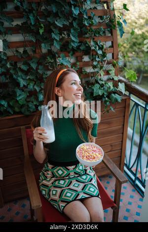Une jeune femme caucasienne joyeuse aux cheveux rouges, inspirée par la mode des années 1950, est assise sur un balcon entouré de lierre luxuriant. Elle tient une bouteille de lait et un bo Banque D'Images