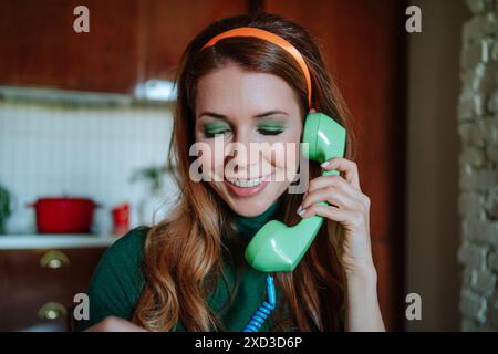 « Une jeune femme rousse dans une cuisine sur le thème des années 1950, s'engageant de manière ludique dans une conversation téléphonique à l'aide d'un téléphone rotatif vert vintage. Elle sourit délicatement, Banque D'Images