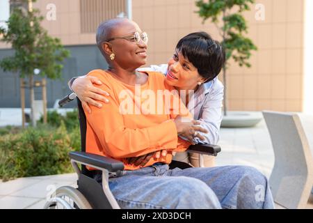 Une femme afro-américaine en fauteuil roulant partage un moment tendre avec une femme latine en plein air, véhiculant amitié et attention Banque D'Images