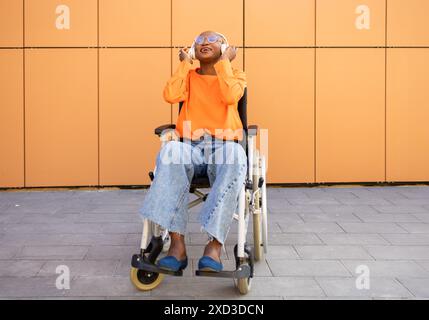 Une femme afro-américaine souriante dans une élégante chemise orange et un Jean, assise dans un fauteuil roulant, écoutant de la musique Banque D'Images