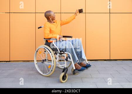 Une femme d'affaires afro-américaine en fauteuil roulant capture un selfie sur un fond orange moderne, mettant en valeur l'indépendance et l'étreinte technologique Banque D'Images