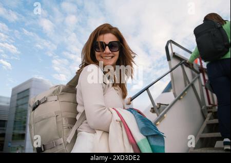 Une femme joyeuse portant des lunettes de soleil et portant un sac à dos monte les escaliers à l'extérieur de l'avion, exsudant un sens de l'aventure. Banque D'Images