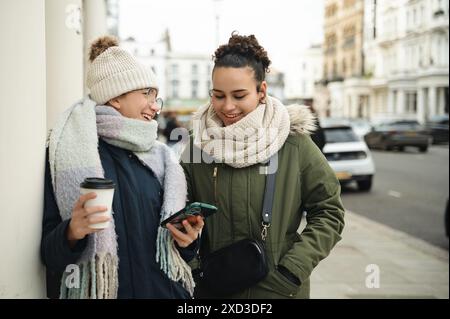 Deux jeunes femmes échangent des sourires tout en s’engageant avec des smartphones dans une rue londonienne dynamique, des cafés chauds à la main, mettant en valeur la connectivité urbaine. Banque D'Images