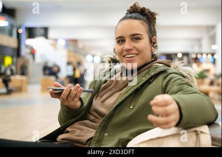 Une jeune femme heureuse regardant la caméra tout en utilisant un smartphone dans un environnement intérieur animé, la foule floue en arrière-plan. Banque D'Images
