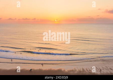 Un coucher de soleil tranquille baigne les surfeurs et la plage dans une lumière chaude, capturant l'essence des paysages d'été sur la côte. Banque D'Images