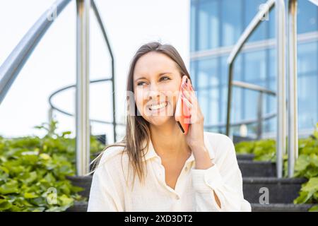 Une jeune femme joyeuse dans une blouse blanche utilise un smartphone tout en étant assise sur les marches à l'extérieur d'un bâtiment moderne en verre Banque D'Images
