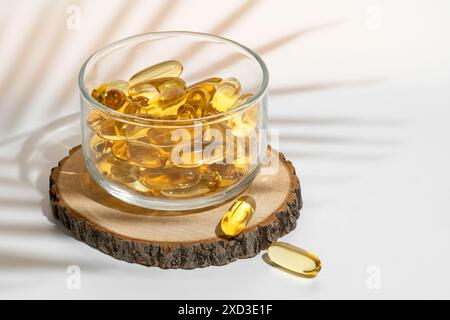 Un bol en verre transparent rempli de capsules oméga 3 dorées est placé sur une tranche de bois rustique, mis en évidence par une lumière douce et diffuse en arrière-plan, créa Banque D'Images