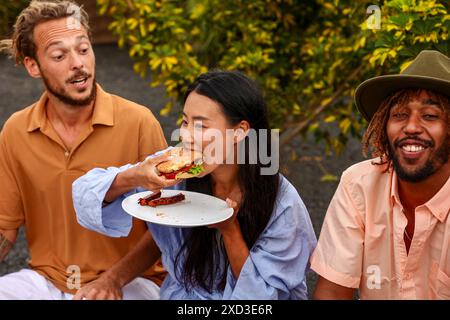 Un groupe multiethnique d'amis profite d'un barbecue à l'extérieur. Une femme asiatique mord dans un hamburger, tandis qu'un homme caucasien et un homme afro-américain conversent, Banque D'Images
