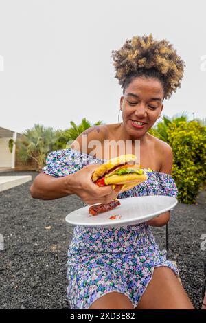 Une jeune femme afro-américaine est assise à l'extérieur, souriant joyeusement tout en regardant vers le bas un copieux hamburger. Vêtue d'une robe d'été fleurie, elle aime Banque D'Images