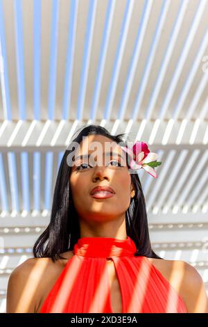 Une femme élégante porte une robe rouge éclatante avec une fleur délicate nichée dans ses cheveux, debout sur un fond rayé moderne Banque D'Images