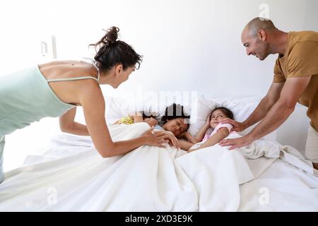 Une famille multiethnique se réveille ensemble dans le lit les parents interagissent amoureusement avec leurs trois enfants, qui sont sous des couvertures blanches brillantes, joyeux matin Banque D'Images