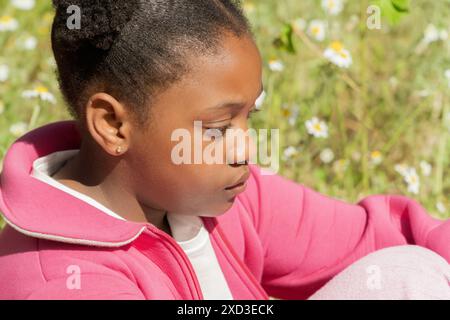 Une jeune fille portant un sweat à capuche rose est capturée dans la contemplation alors qu'elle est assise à l'extérieur, entourée de fleurs sauvages dans un cadre de parc animé Banque D'Images