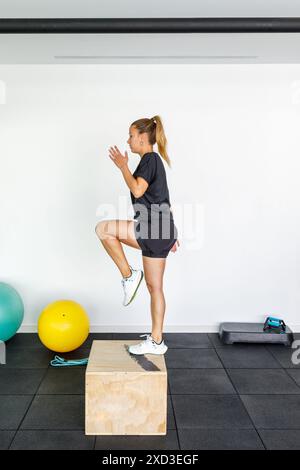 Une jeune femme en tenue sportive effectue un exercice de saut de boîte dynamique dans un gymnase contemporain et bien équipé, en se concentrant sur le renforcement de la force et de l'agili Banque D'Images