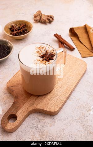 D'en haut, délicieux chai latte avec de la mousse crémeuse sur une planche de bois entourée d'anis étoilé, de bâtonnets de cannelle, de gingembre frais, de feuilles de thé, d'un pot de lait A. Banque D'Images