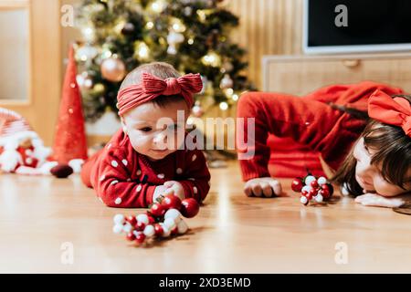 Deux jeunes frères et sœurs dans des tenues rouges festives profitent d'un moment ludique sur un parquet pendant Noël, entourés de décorations de vacances. Banque D'Images