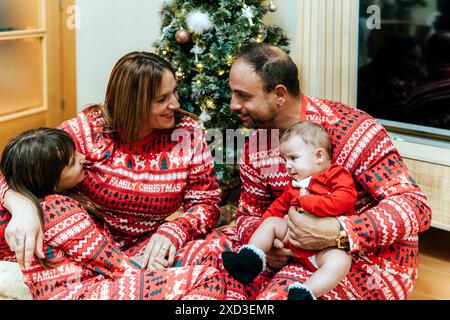 Une famille heureuse avec deux adultes et deux enfants portant un pyjama de Noël festif souriant et profitant de la compagnie de l'autre près du sapin de Noël. Banque D'Images