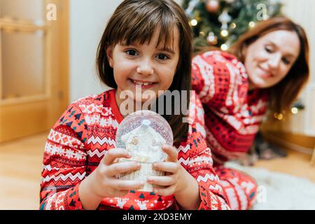 Une petite fille, regardant la caméra avec plaisir à un globe de neige, est accompagnée par une mère souriante la regardant, les deux portant une sueur de Noël festive Banque D'Images