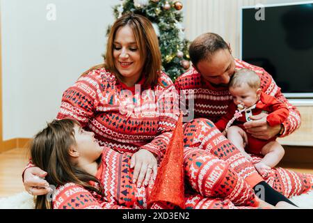 Une famille de quatre personnes portant un pyjama de Noël Red Family assorti est assis près de leur sapin de Noël décoré, se liant à un bébé. Banque D'Images