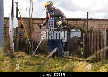 Un homme en équipement de sécurité garnit de grandes herbes sauvages dans une cour encombrée avec un coupe-ficelle la scène comprend divers outils de jardinage et un bois rustique Banque D'Images