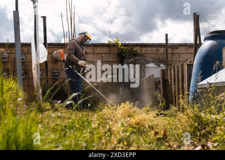 Un homme en équipement de protection utilise un coupe-herbe pour nettoyer l'herbe envahie dans une cour arrière négligée, mettant en valeur la tâche de l'entretien du jardin Banque D'Images