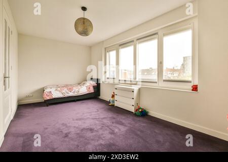 Une chambre avec tapis violet et murs blancs, il y a un petit lit d'enfant devant la fenêtre Banque D'Images