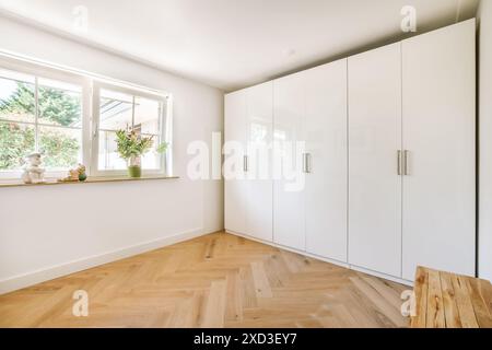 Cette chambre contemporaine dispose d'élégantes armoires encastrées blanches, de parquet lumineux et d'une grande fenêtre laissant passer la lumière naturelle. Parfait pour le mode Banque D'Images