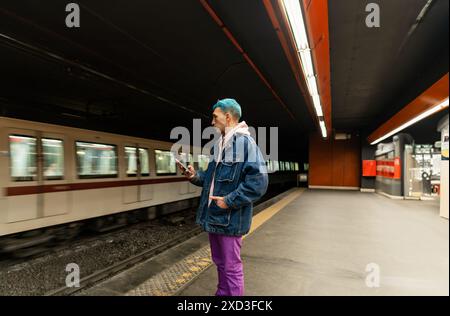 Un jeune homme aux cheveux bleus éclatants se tient sur une plate-forme de métro urbain, vérifiant son téléphone, alors qu'un train roule en arrière-plan Banque D'Images