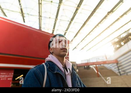 Un jeune homme à la mode portant des lunettes de soleil roses et une veste en Jean se tient en toute confiance devant une gare, entouré d'un architecte contemporain saisissant Banque D'Images