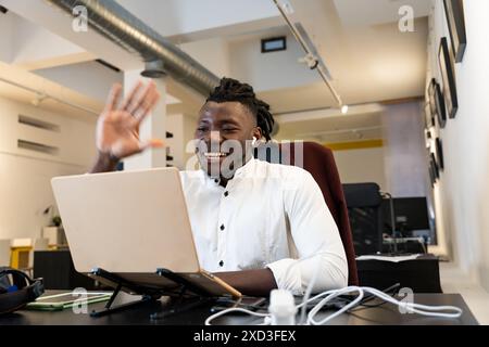 Un jeune homme joyeux dans une chemise blanche est engagé dans un appel vidéo, agitant joyeusement à l'écran depuis son espace de travail organisé Banque D'Images