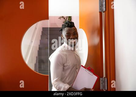 Un jeune homme professionnel et joyeux se tient près d'une porte de bureau ouverte, tenant un presse-papiers, souriant doucement vers la caméra d'une manière invitante Banque D'Images