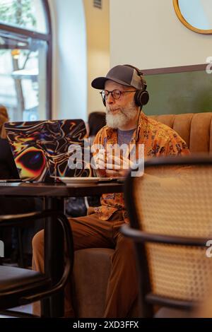Un homme âgé moderne dans une chemise vibrante et casquette travaillant à distance sur un ordinateur portable à motifs, avec des écouteurs dans un cadre de café confortable Banque D'Images