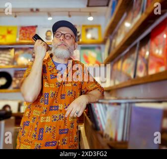 Homme senior avec barbe et lunettes, portant une chemise colorée et une casquette, feuilletant soigneusement les disques vinyles dans un magasin de musique confortable Banque D'Images