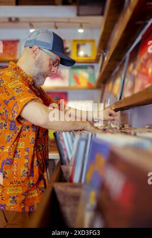 Un homme âgé actif dans une chemise vibrante et une casquette de baseball est engagé dans la sélection de disques vinyles dans un magasin de musique, mettant en valeur un style de vie senior moderne Banque D'Images