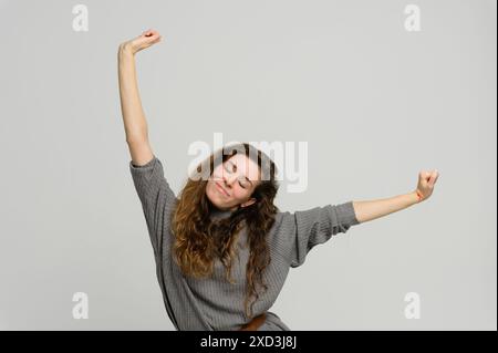 Une jeune femme attirante avec de longs cheveux s'étire, levant les bras au-dessus de sa tête. Femme se réveillant en étirant les bras. Fond blanc. Banque D'Images