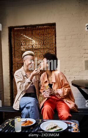Un couple diversifié de lesbiennes profitant d'un repas ensemble sur un banc. Banque D'Images