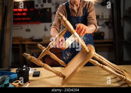 Homme portant des gants orange et un tablier réparant une vieille chaise en bois dans un atelier. Il enroule du ruban adhésif autour des pieds de la chaise avant de peindre. Banque D'Images