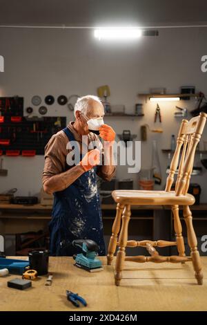 Un homme âgé en gants orange, un tablier bleu, se tient dans l'atelier devant une vieille chaise en bois, met un masque de protection sur son visage. Banque D'Images