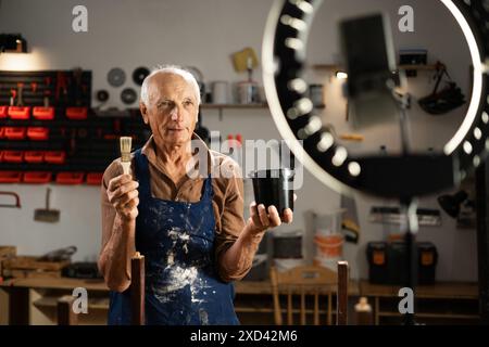 homme dans un tablier et des gants orange enregistre vlog ou streaming en direct sur la restauration de meubles dans son atelier. Banque D'Images