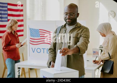 Jeune citoyen noir confiant plaçant un bulletin de vote dans l'urne au lieu de vote le jour de l'élection présidentielle Banque D'Images