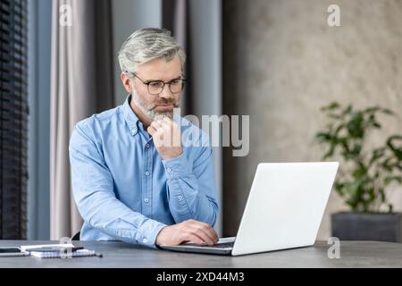 Homme mûr avec des lunettes assis au bureau, profondément concentré sur son travail à l'aide d'un ordinateur portable. Environnement de bureau moderne. Banque D'Images