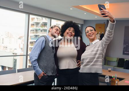 des collègues, deux femmes et un homme, des amis d'ethnie différente, sont debout en train de prendre un selfie au bureau avec un téléphone portable, l'une des femmes a Banque D'Images